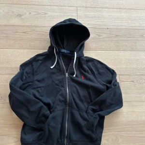 Svart hoodie från Polo Ralph Lauren - Säljer en svart hoodie från Polo Ralph Lauren med dragkedja och en liten röd broderad logga på bröstet. Den har en justerbar huva med vita snören och är perfekt för en avslappnad stil. Passar bra till både jeans och joggers. Den har en liten defekt vid nacken, se 4e bilden. Sitter som en Medium