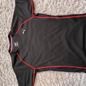 Svart tränings t-shirt från Puma - Säljer en svart tränings t-shirt från Puma med röda sömmar och korta ärmar. Perfekt för träning med sitt lätta och bekväma material. Puma-loggan syns på bröstet. Passar bra för en aktiv livsstil.