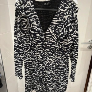 Svart och vit mönstrad klänning från Zara - Snygg svart och vit klänning med ett unikt mönster. Klänningen har en v-ringad design och långa ärmar, perfekt för en stilfull look. Passar bra till både vardag och speciella tillfällen.
