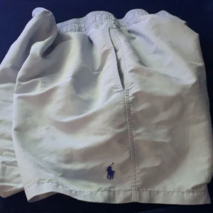 Bad shorts från Ralph Lauren. Perfekt för sommaren som kommer nu - Snygga beige shorts från Ralph Lauren med elastisk midja och en liten broderad logga i blått. Perfekta för en avslappnad stil. De har en bakficka med knapp för extra säkerhet. 