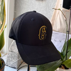 Svart keps från Yupoong - Snygg svart keps från Yupoong med Flexfit-teknologi. Kepsen har en gul broderad logga framtill och är tillverkad i polyester. Perfekt för en casual look.