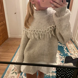 Beige stickad tröja med fransdetaljer - Säljer en mysig beige stickad tröja med unika fransdetaljer över bröstet. Tröjan har långa ärmar och en ribbad krage som ger en stilren look. Perfekt för kyliga dagar!