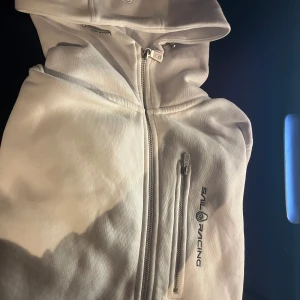 Sail racing - Säljer en stilren vit huvtröja från Sail Racing med dragkedja och broderad logga på bröstet. Tröjan har en praktisk huva och en ficka med dragkedja framtill. Perfekt för en avs