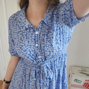 Blå blommig klänning - Blå klänning med blommigt mönster och band som går att knyta i midjan som framhäver midjan. Den har korta ärmar och knappar framtill, perfekt för en sommaren💙 Inte min stil så säljer, vid intresse är det bara att skriva😊💕