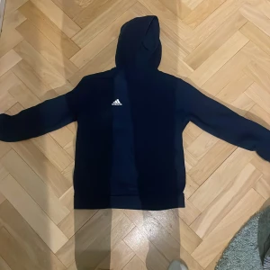 Mörkblå hoodie från Adidas - Säljer en snygg mörkblå hoodie från Adidas med klassisk logga på bröstet. Perfekt för en avslappnad stil med sin bekväma passform och huva. Passar bra till både träning och vardag. 🌀