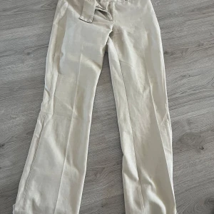 Beige kostymbyxor - Snygga beige kostymbyxor med mid midja och raka ben. Perfekta för en stilren look. Byxorna har en klassisk design med knappar och dragkedja framtill. Lite mindre i stl