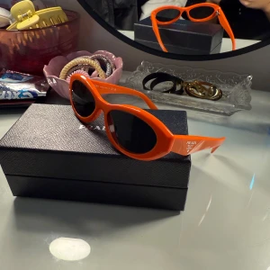 Orange solglasögon från Prada - Snygga orange solglasögon från Prada med en modern design. De har svarta linser och en stilren båge som ger en cool look. Perfekta för att sticka ut i mängden och skydda ögonen med stil. Knappt använda!! Finns både kvitto och original förpackning!