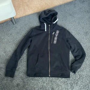 Svart hoodie med dragkedja - Säljer en svart hoodie med dragkedja och tryck på bröstet. Den har en justerbar huva med vita snören och praktiska fickor framtill. Perfekt för en avslappnad stil. 