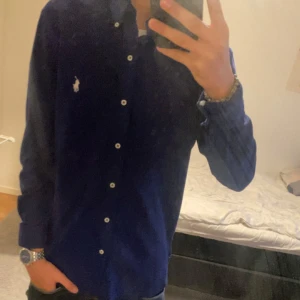 Mörkblå skjorta från Ralph Lauren - Snygg mörkblå skjorta från Ralph Lauren med klassisk krage och broderad logga på bröstet. Skjortan har långa ärmar och knappar framtill. Perfekt för en stilren look.