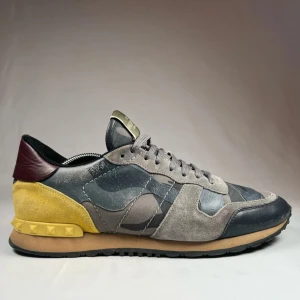 Valentino Rockrunner i grått och gult - Snygga Valentino Rockrunner sneakers i en unik kombination av grått, gult och vinrött. Skorna har ett coolt camouflagemönster och är tillverkade i en mix av mocka och skinn. De har snörning och en bekväm sula för optimal komfort.