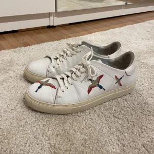 Vita sneakers från Axel Arigato - Säljer nu ett par sjukt snygga vita sneakers från Axel Arigato med färgglada fåglar på sidorna. Skorna har snörning och är bekväma att gå i så dom funkar perfekt till vardags. Dom är sparsamt använda och köpta för ca 2800 i deras butik. Lite smutsiga men annars i väldigt bra skick💕