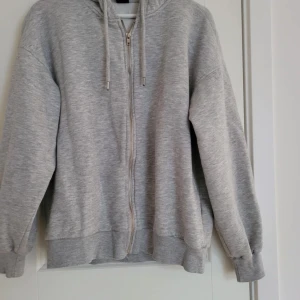 Grå hoodie med dragkedja - Säljer en bekväm grå hoodie med dragkedja och justerbar huva. Perfekt för en avslappnad stil. Den har långa ärmar och ribbade muddar vid ärmslut och nederkant.