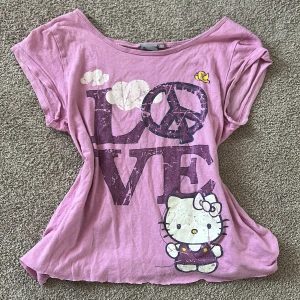 Rosa Hello Kitty t-shirt - Säljer en söt rosa t-shirt med Hello Kitty-tryck och texten 'LOVE' i stort lila tryck. T-shirten har korta ärmar och offshoulder axlar. Den är köpt direkt från en sanrio butik. Jag (tror) inte att den går att köpa i butik idag. Den är egentligen storlek 170 men jag skulle säga att den är onesize eftersom att den passar mig som är 164💕💕Jag har själv klippt upp den eftersom att den var vääldigt lång innan !!