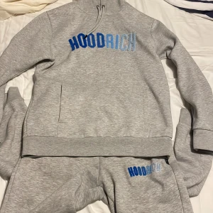 Grå dress från Hoodrich - Säljer en grå hoodie från Hoodrich med blå broderad text på bröstet. Tröjan har en dragkedja vid halsen och ribbade muddar vid ärmslut och nederkant. Perfekt för en avslappnad stil.