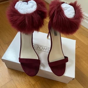 Vinröda klackskor med pälsdetalj - Snygga vinröda klackskor med pälsdetalj runt ankeln. Skorna har en öppen tå och en elegant klack, perfekt för att sticka ut på festen. Använda en gång. Fortfarande i original pack