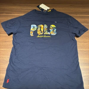 Mörkblå t-shirt från Ralph Lauren - Säljer en snygg mörkblå t-shirt från Ralph Lauren med ett färgglatt tryck av ordet 'POLO' på framsidan. Spontan köpte t-shirten i Ungern för 40k HUF vilken motsvarar 1100kr. Den är ALDRIG använd. Har du några fråger eller vill ha mer bilder så är det bara attt skriva till mig här på plick 🤗