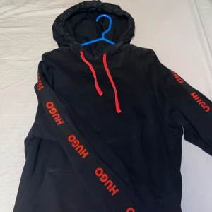 Svart hoodie från Hugo Boss - Hej Säljer en svart hoodie från Hugo Boss med röda detaljer. Den har en stor ficka framtill och röda snören i huvan. Ärmarna pryds av Hugo-loggan i rött. Perfekt för en casual look!