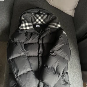 Svart pufferjacka med rutigt foder - Säljer en svart pufferjacka med ett snyggt rutigt foder i grått och vitt. Jackan har en dragkedja framtill och långa ärmar. Perfekt för kyliga dagar!