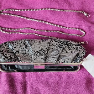 Svart och silver broderad kuvertväska - Elegant kuvertväska i svart och silver med vackert broderat mönster. Väskan har en avtagbar kedja i silverfärg som gör den lätt att bära över axeln. Perfekt för att ge en lyxig touch till din outfit. 25 cm bred. 