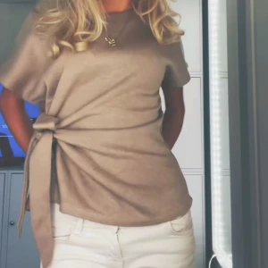 Beige knyttopp - Säljer en stilren beige topp med knytning i sidan. Toppen har korta ärmar och en mjuk, bekväm passform. Perfekt för en avslappnad men chic look.