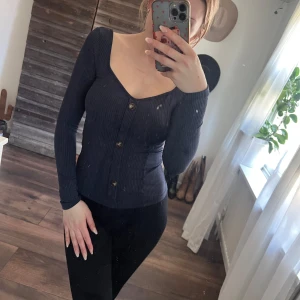Svart ribbad topp - Snygg svart ribbad topp med knappar framtill. Den har en v-ringad design och långa ärmar, perfekt för en stilren look. Passar bra till både jeans och kjol.