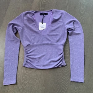 Lila glittrig topp från Bik Bok - Säljer en snygg lila glittrig topp från Bik Bok. Toppen har en v-ringad design och långa ärmar. Den är figurnära med rynkade detaljer i midjan som ger en smickrande passform. Perfekt för att lysa upp vilken outfit som helst!