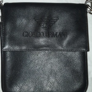 Svart axelväska från Giorgio Armani - Snygg svart axelväska från Giorgio Armani i skinn. Väskan har en stilren design med en praktisk dragkedja på baksidan och en justerbar axelrem i metall. Perfekt för att bära dina viktigaste saker med stil.