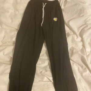 Svarta mjukisbyxor med blomdetalj - Säljer ett par svarta mjukisbyxor med en söt blomdetalj på ena benet. Byxorna har en bekväm passform med dragsko i midjan för justerbarhet.