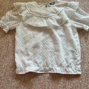 Vit blus från WE - Säljer en söt vit blus från WE med volangdetaljer och korta ärmar. Blusen har ett subtilt mönster och en knapp i nacken för enkel påklädning. Perfekt för en stilren look!