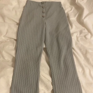 Randiga byxor med knappar - Snygga randiga byxor i en grå nyans med knappar framtill. Byxorna har en tight passform och är perfekta för en stilren look. De är högmidjade och har ett klassiskt mönster som ger en tidlös känsla.