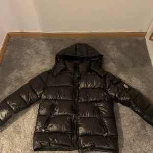 Svart dunjacka från Moncler - Säljer en snygg svart dunjacka från Moncler med huva och dragkedja. !Fickan på vänstra sidaan är sönder på insidan, men inget man ser!Jackan har en glansig finish och två praktiska fickor med dragkedjor. Perfekt för kyliga dagar när du vill hålla stilen. 🖤