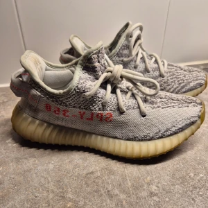 Adidas Yeezy Boost 350 V2 - Säljer ett par Adidas Yeezy Boost 350 V2 Beluga i grått med den ikoniska texten 'SPLY-350' i rött på sidan. Skorna har en stickad ovandel och en ribbad sula i beige. Perfekta för dig som vill ha en stilren och bekväm sneaker.
