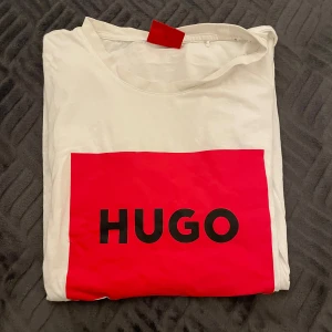 Vit t-shirt från Hugo - Säljer en stilren vit t-shirt från Hugo med en stor röd logga på framsidan. Perfekt för en casual look med en touch av stil. T-shirten har korta ärmar och är gjord i mjukt material.