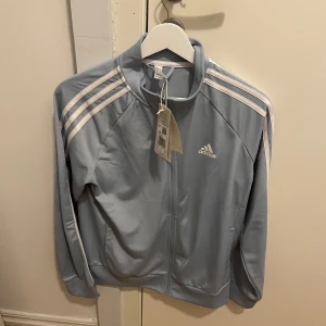 Ljusblå träningsjacka från Adidas - Säljer en snygg ljusblå träningsjacka från Adidas med de klassiska tre ränderna på ärmarna. Jackan har dragkedja framtill och en bekväm passform. Perfekt för träning eller en sportig look. 💙