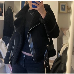Svart teddyjacka med dragkedja - Säljer en snygg svart teddyjacka med dragkedja och skinnliknande detaljer. Jackan har en cool bikerstil med en asymmetrisk dragkedja och långa ärmar. Perfekt för att hålla sig varm och stilren under kyliga dagar.