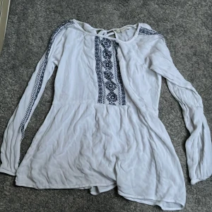 Vit blus från Happy Holly - Säljer en vit blus från Happy Holly med blå broderade detaljer längs ärmarna och framtill. Blusen har långa ärmar och knytning vid halsen. Perfekt för en bohemisk stil.