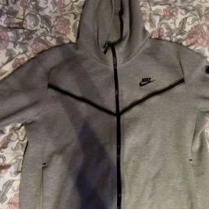 Grå huvtröja från Nike - Säljer en grå huvtröja från Nike med dragkedja och svart logotyp på bröstet. Tröjan har långa ärmar och en stilren design med svarta detaljer längs dragkedjan och ärmarna. Perfekt för en sportig look.