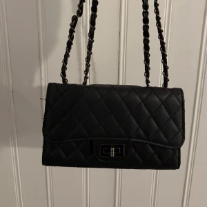 Svart quiltad axelväska - Snygg svart quiltad axelväska med kedjehandtag och elegant låsdetalj framtill. Perfekt för att ge en stilren touch till din outfit. 🖤
