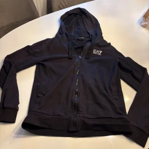 Svart hoodie från Emporio Armani storlek S - Säljer en stilren svart hoodie från Emporio Armani med dragkedja och huva. Perfekt för en avslappnad look. Den har långa ärmar och en diskret logga på bröstet.