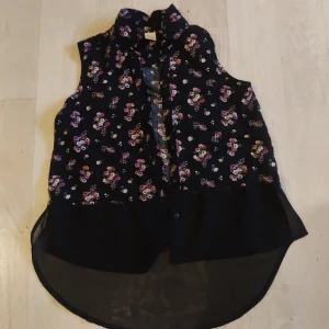 Blommig ärmlös blus från Lindex - Säljer en söt blommig ärmlös blus från Lindex i svart med små rosa och vita blommor. Perfekt för varma dagar med sin lätta och luftiga design. Blusen har en längre bakdel och knappar framtill.