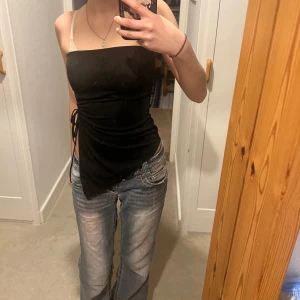 Svart axelbandslös topp - Snygg svart axelbandslös topp med knytning i sidan och en cool cut-out detalj. Perfekt för en kväll ute eller när du vill känna dig extra chic. Toppen har en tight passform som framhäver figuren.