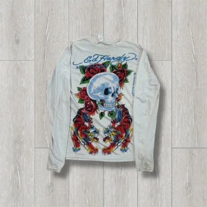 Vit långärmad tröja från Ed Hardy - Säljer en cool vit långärmad tröja från Ed Hardy med ett stort tryck av en dödskalle, rosor och tigrar i färgglada nyanser. Tröjan har blå textdetaljer och är perfekt för dig som gillar att sticka ut. Passar bra till både jeans och kjol. 🐯🌹Tröjan har små hål som har sytts och lite små som är osydda men ändå bra skick