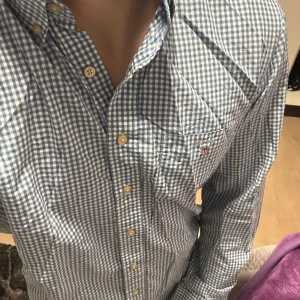 Blårutig skjorta från GANT - Snygg blårutig skjorta från GANT i storlek M. Skjortan har långa ärmar och klassiska knappar framtill. Perfekt för en stilren look med en liten broderad logga på bröstet. Passar utmärkt till både vardag och mer uppklädda tillfällen.