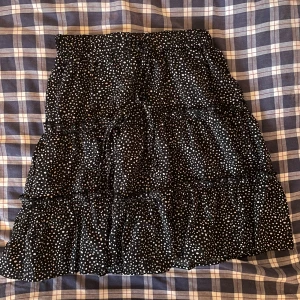 Svart prickig kjol - Snygg svart kjol med vita prickar och volanger. Perfekt för en trendig look. Resår i midjan för bekväm passform.