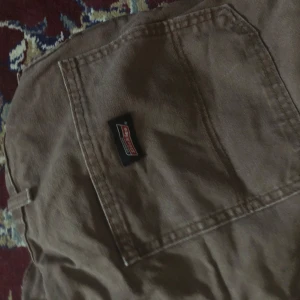 Bruna byxor från Dickies - Säljer ett par bruna byxor från Dickies. De har en klassisk design med en stor ficka på sidan och är perfekta för en avslappnad stil. Byxorna är tillverkade i ett slitstarkt material och har en oversized passform