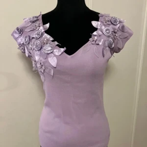 Ljuslila topp med super fina blommiga detaljer - Säljer en unik ljuslila topp med v-ringning och korta ärmar. Toppen har vackra blommiga applikationer och blad i satin på axlarna, vilket ger en romantisk touch. Perfekt för att sticka ut på speciella tillfällen. Helt ny, har aldrig kommit till användning.