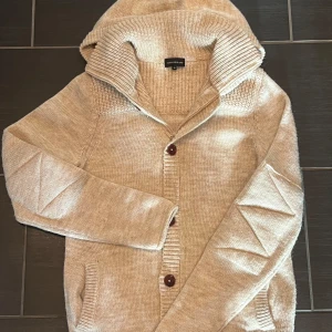 Beige cardigan - Tja! Säljer denna sjukt snygga och unika cardiganen. Cardiganen har en huva och knappar framtill. Riktigt detaljerad kofta med snygga detaljer! Cardiganen är storlek S men skulle säga att den även passar storlek M. Hör av dig vid minsta fråga!