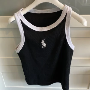 Svart linnetopp från Ralph Lauren - Snygg svart linnetopp från Ralph Lauren med vita kanter och det klassiska broderade logotypen på bröstet. Perfekt för en stilren och avslappnad look. Köpt utomlands så osäker på äktheten, men mycket snygg!🙌💫