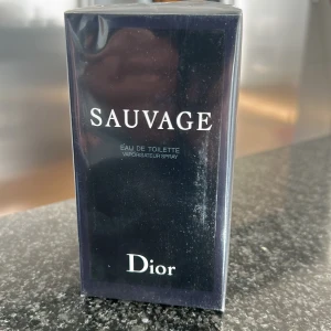 Dior Sauvage Eau de Toilette - Säljer en elegant flaska av Sauvage Eau de Toilette från Dior. Flaskan har en stilren design i mörkblått med vita detaljer. Perfekt för den som söker en tidlös och maskulin doft. Flaskan är förseglad och redo att användas. Oöppnad 100 ml