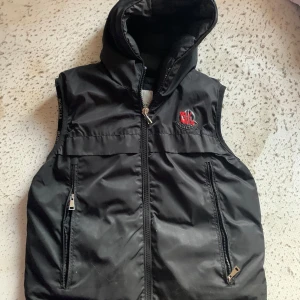 Moncler x Palm angels  - Säljer en snygg svart dunväst från Moncler med huva och dragkedja. Västen har två fickor med dragkedjor och ett broderat Moncler-märke på bröstet. Perfekt för kyliga dagar!